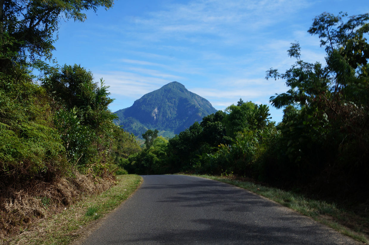 A narrow, paved road crests a hill with green vegetation on either side and a green, triangular mountain in the distance.